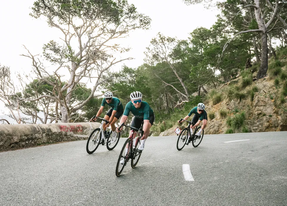 Drie fietsers in groene truien en witte helmen fietsen op een weg in een heuvelachtig landschap.