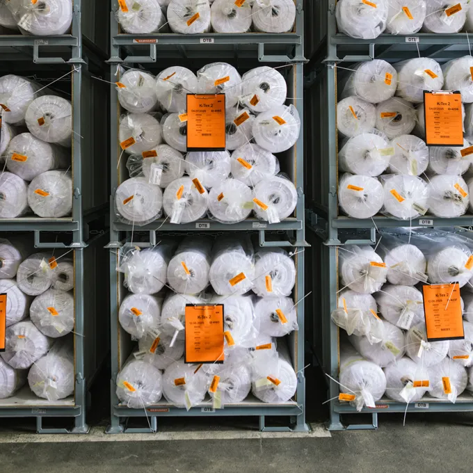 Een magazijn met rekken gevuld met grote rollen witte stof verpakt in plastic met oranje etiketten.