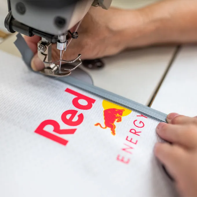 Een close-up van een naaimachine die een wit sportkledingstuk met het Red Bull-logo naait, terwijl iemand het materiaal vasthoudt.