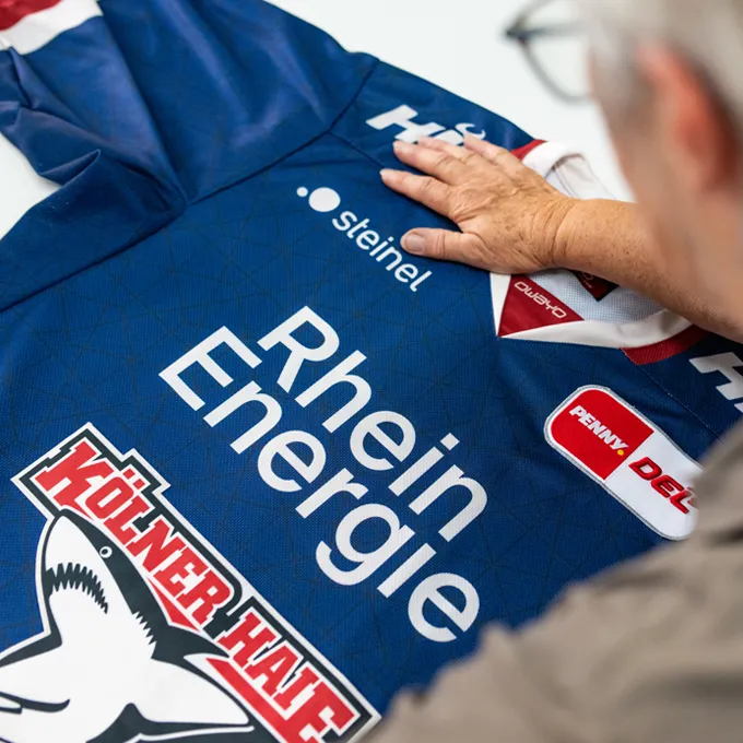 Een persoon controleert een blauw aangepast sportshirt van owayo met verschillende sponsorlogos, waaronder 'Rhein Energie' en 'Kölner Haie'.