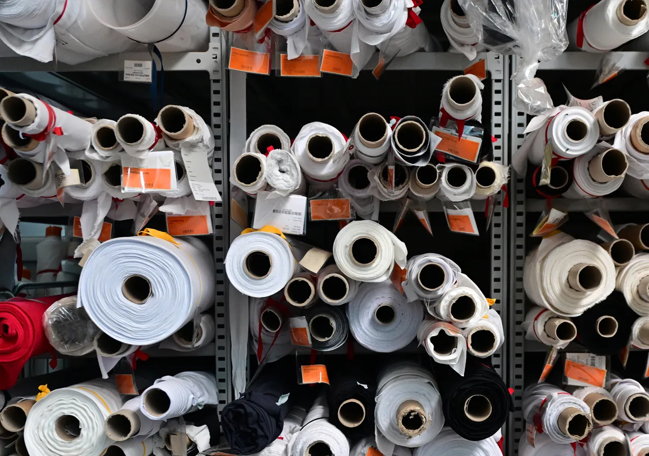 Planken vol met stofrollen in verschillende kleuren, waaronder wit, zwart en rood, met etiketten die wijzen op een eigen productiefabriek.