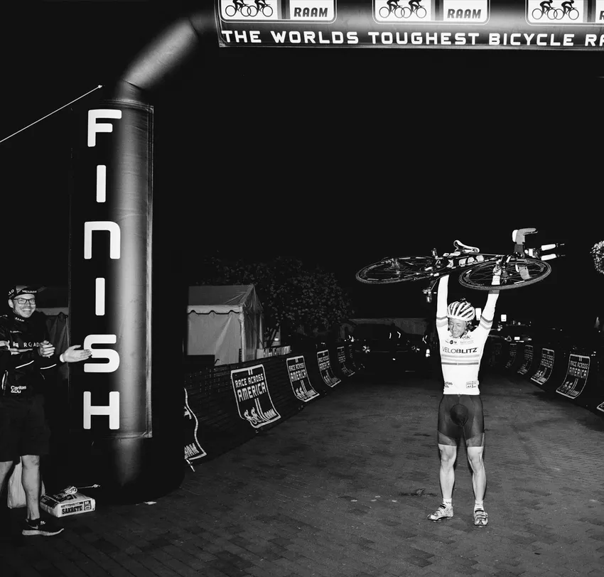 Een wielrenner viert zijn overwinning bij de finish van de 'Race Across America' met zijn fiets boven zijn hoofd, terwijl een man naast hem applaudisseert.