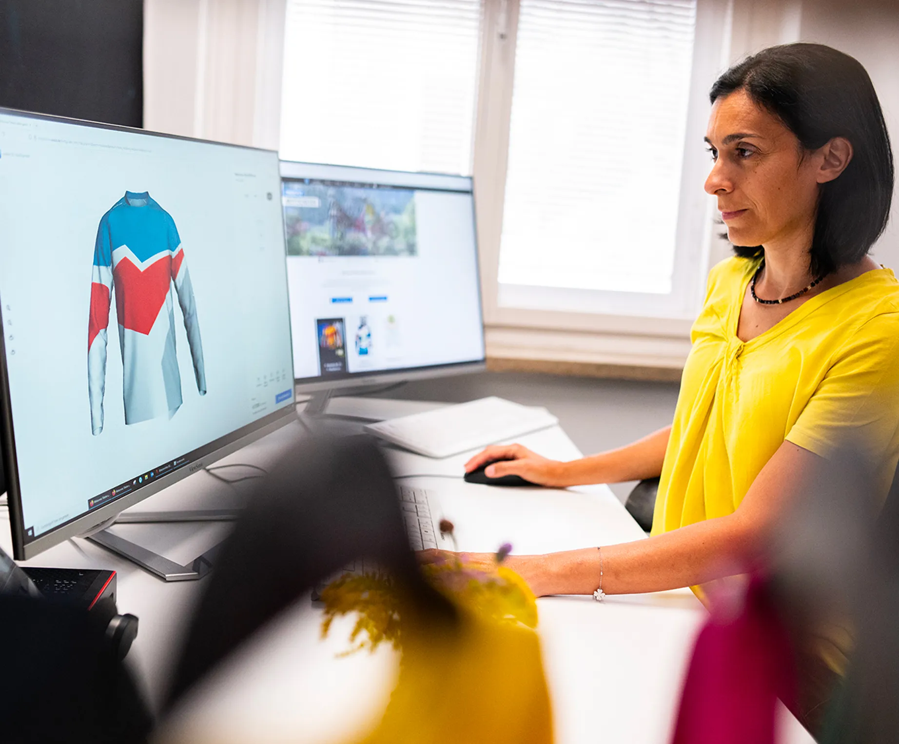 Een vrouw in een geel shirt zit aan een bureau en ontwerpt op een computerbeeldscherm een aangepast sportshirt met blauwe, rode en witte panelen.