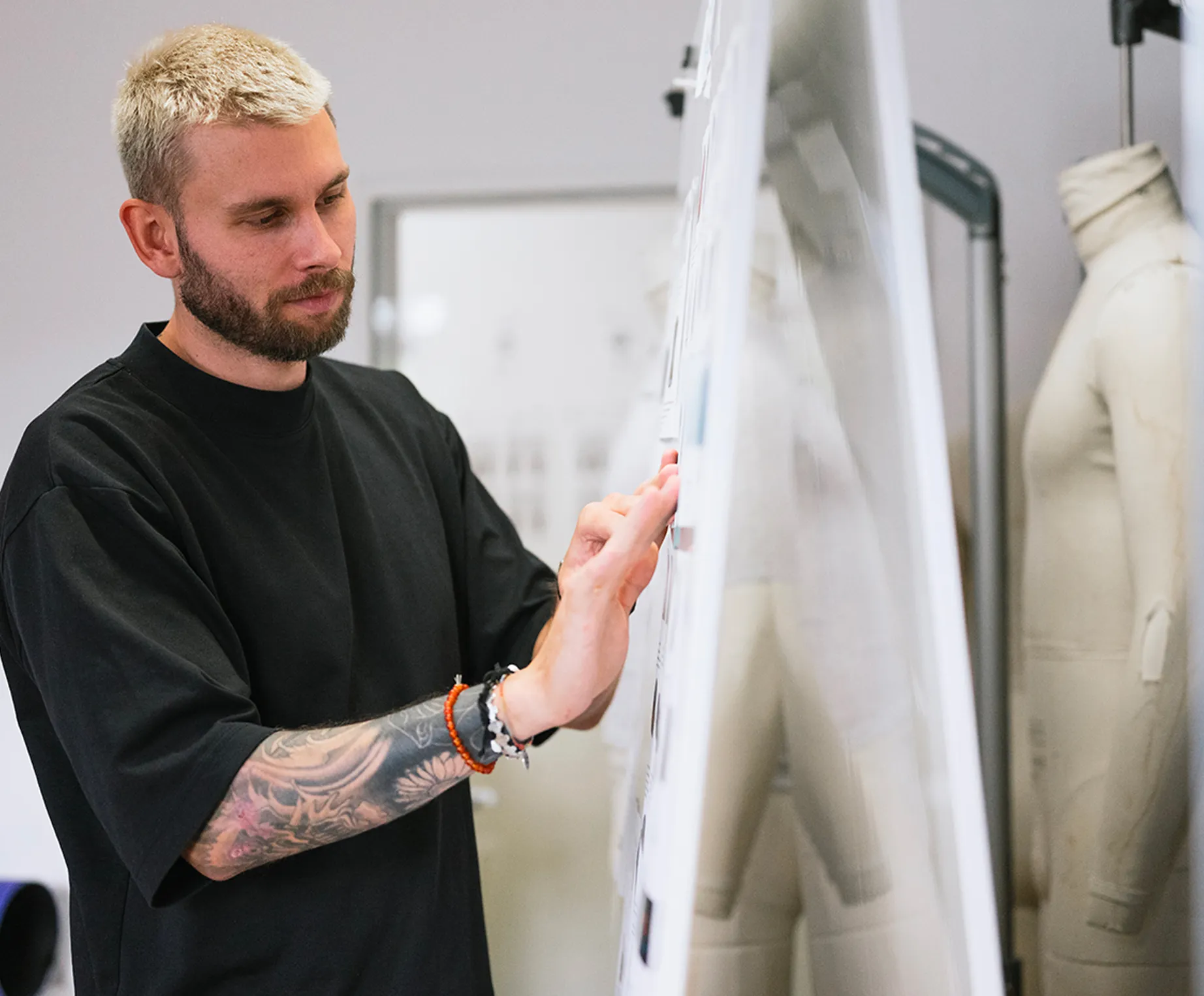 Een man met blond haar en een tatoeage op zijn arm bekijkt geconcentreerd ontwerpen op een prikbord in een ontwerpstudio, terwijl een naaimodel op de achtergrond staat.